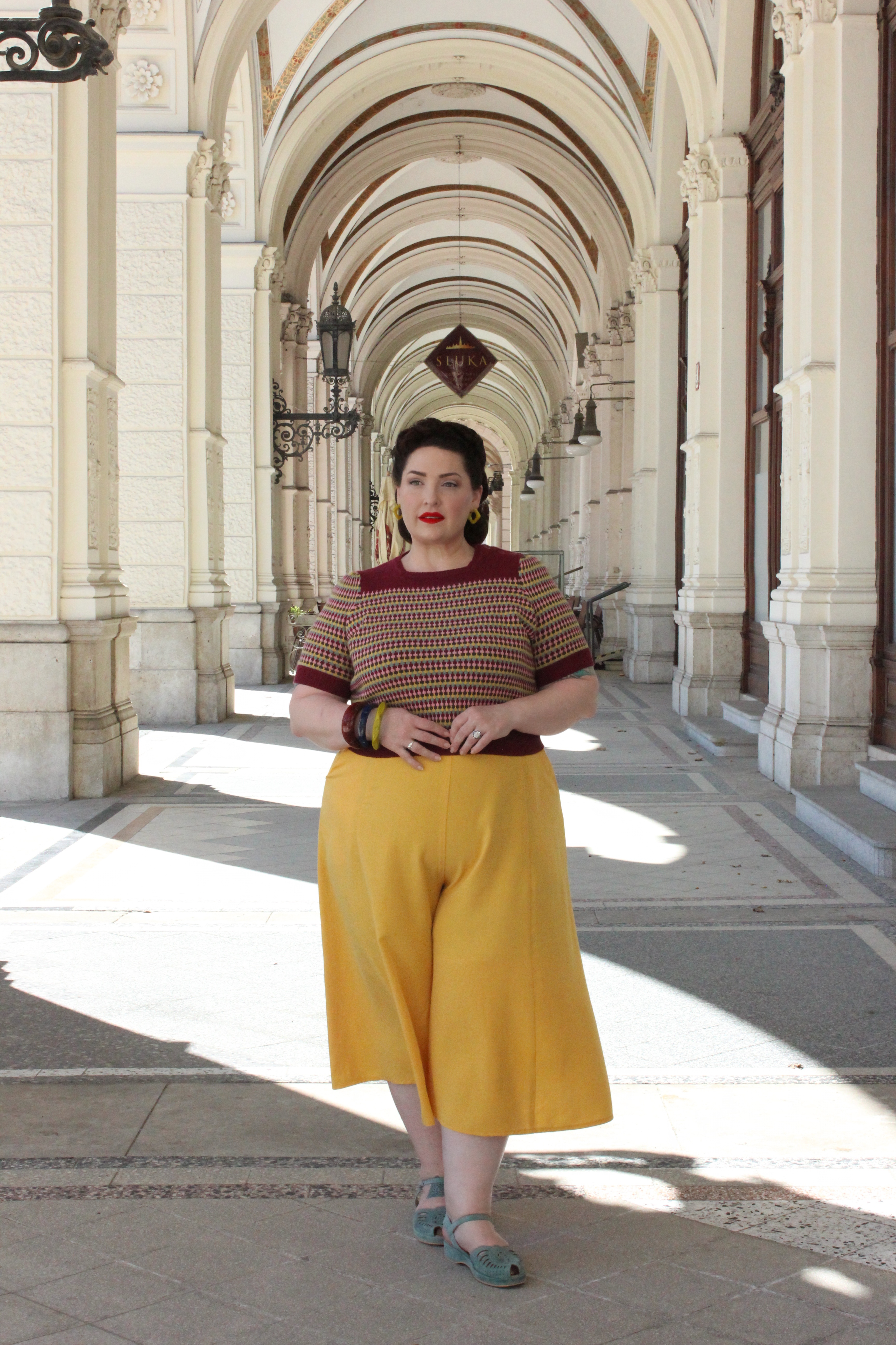 Vintage-inspired spring outfit in Vienna featuring a colorful knit top and culottes, standing under a historic archway